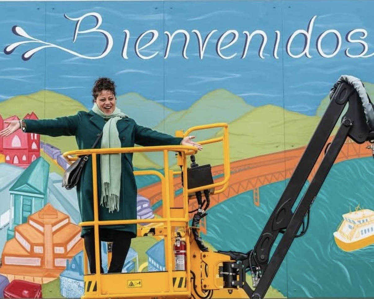An artist on a lift celebrates with open arms in front of a community 'Bienvenidos' mural of the Hudson Valley — rolling hills, a bridge, and a ferry on the water.